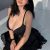 Woman with long dark hair sits on a bed in a black lingerie top and lace-trimmed skirt, smiling at the camera.