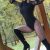 Woman posing on a wooden outdoor frame, wearing a black long-sleeve bodysuit, patterned fishnet tights, knee-high black boots, and a red hat.
